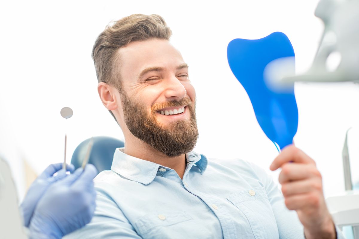 Adult man smiling at his straightened teeth in a hand mirror after orthodontic treatment